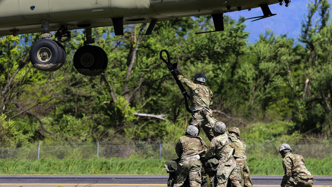 Soldiers stand on a platform trying to hook a line to the bottom of a helicopter.