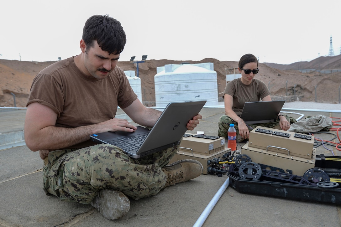 Two service members work on laptops in the desert.