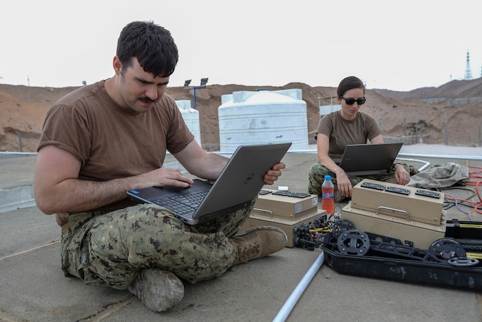 Two service members work on laptops in the desert.