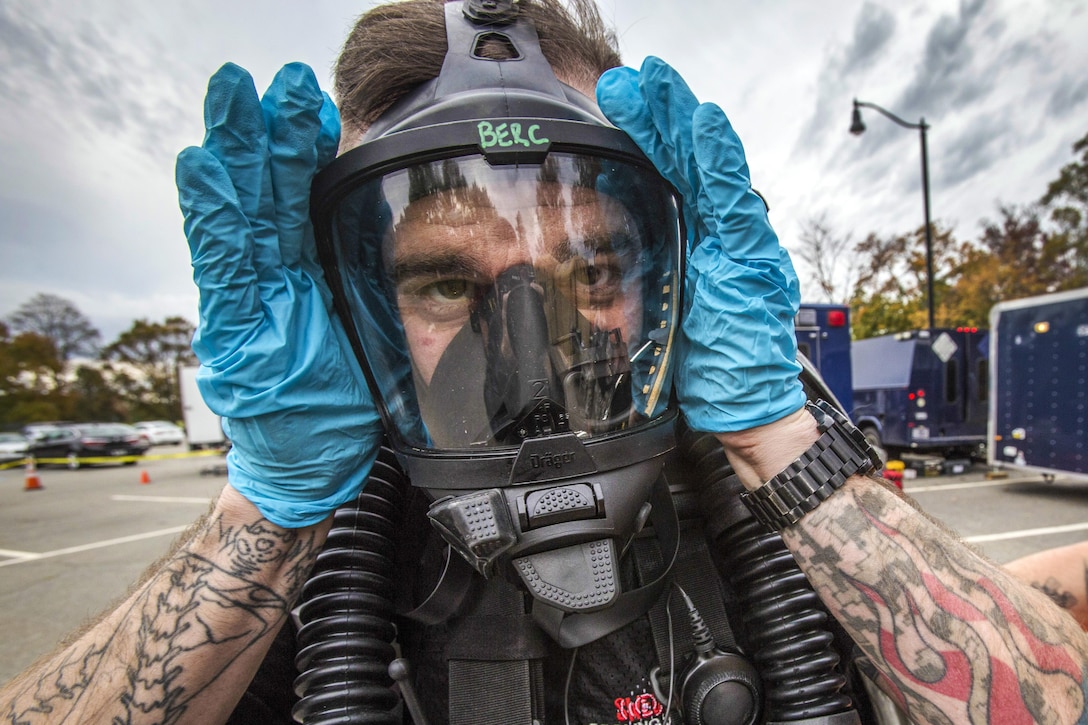 A soldier holds his blue-gloved hands up on either side of a breathing mask he's wearing.
