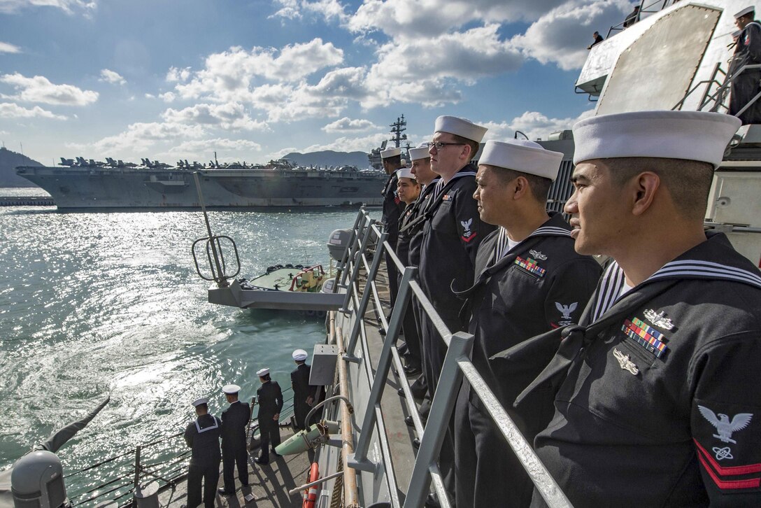 Sailors stand at the rails of a ship coming into port.