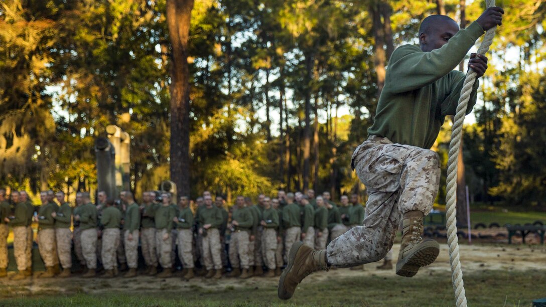 A recruit makes a running leap while holding onto a rope as a crowd of recruits watches in the background.