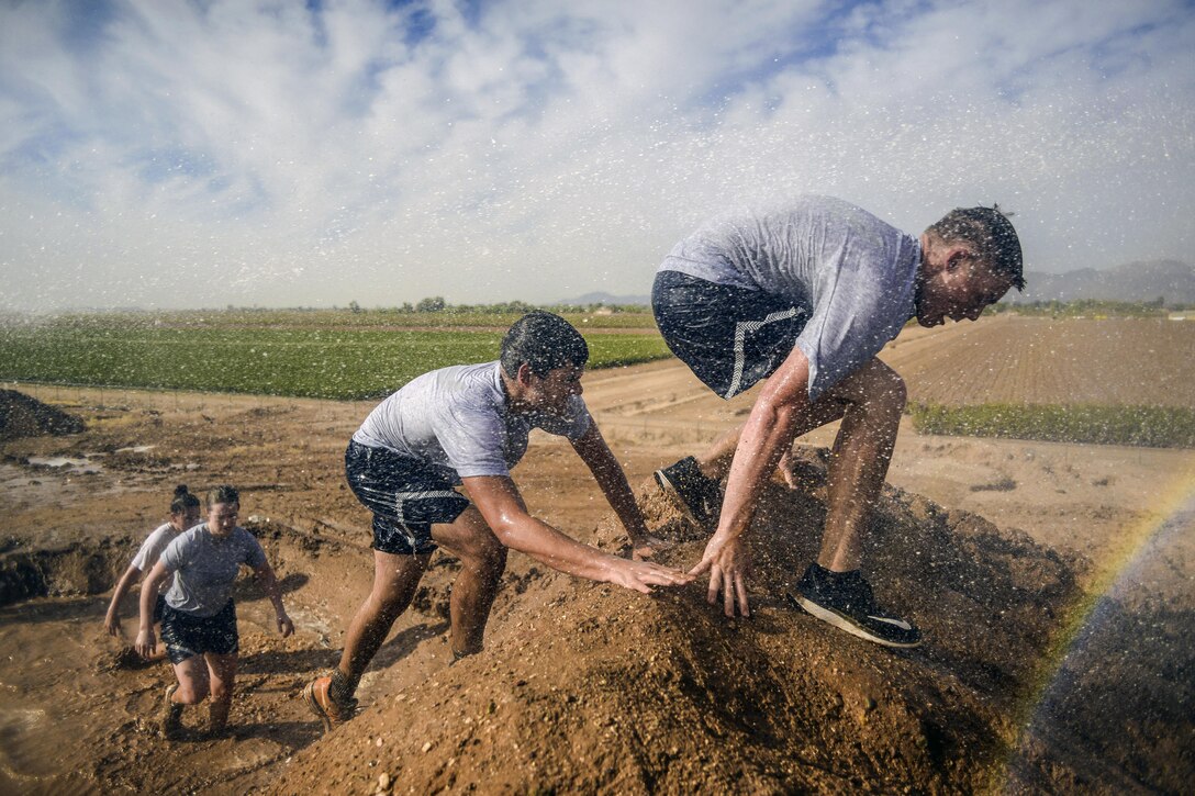 Four cadets work to scale a muddy hill, one behind the other.
