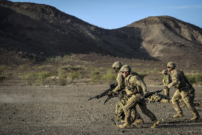 A group of soldiers carrying machine guns rushes forward on flat terrain, with mountains in the background.