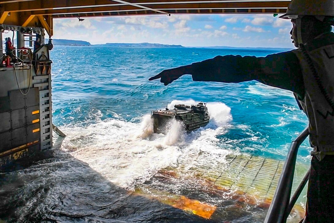 A sailor inside a ship's well deck points, as an amphibious assault vehicle splashes out of the deck and into turquoise water.