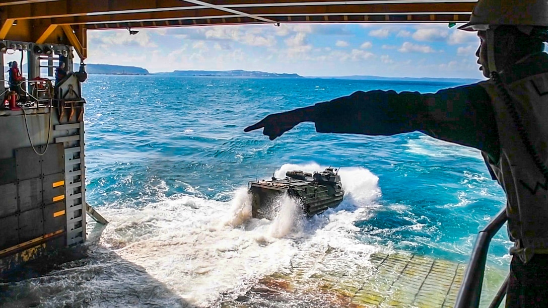 A sailor inside a ship's well deck points, as an amphibious assault vehicle splashes out of the deck and into turquoise water.