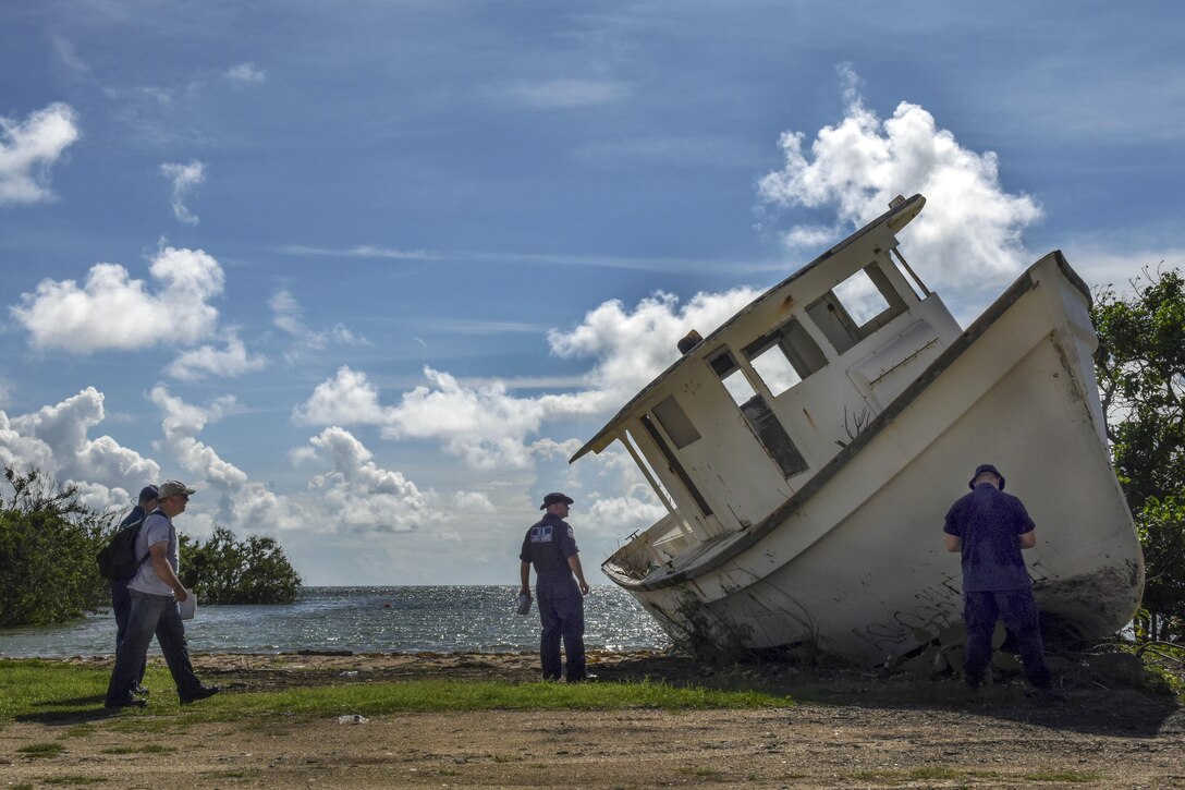 Uniformed personnel examine a muddy boat tipped on its side on a stretch of shore.