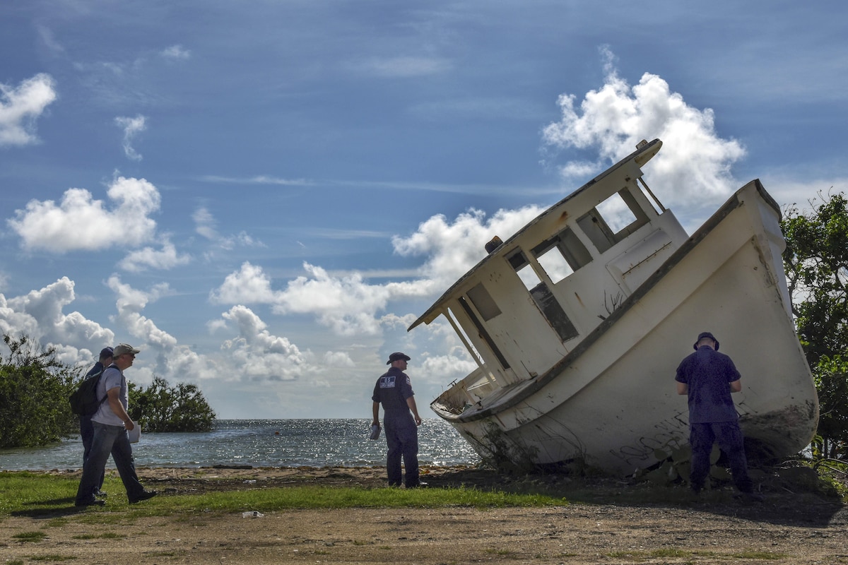 Uniformed personnel examine a muddy boat tipped on its side on a stretch of shore.