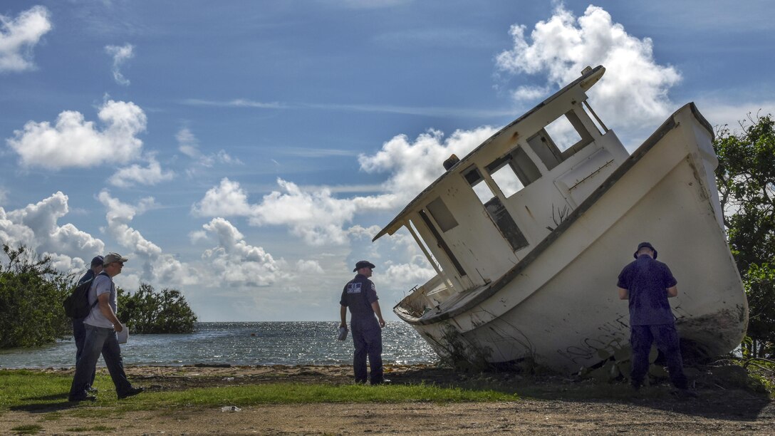 Uniformed personnel examine a muddy boat tipped on its side on a stretch of shore.