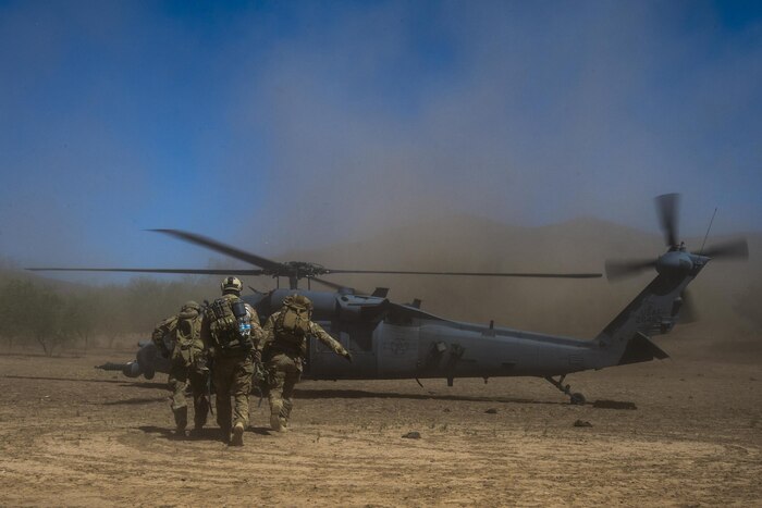 Airmen move a mock patient towards a helicopter that is blowing up dirt.