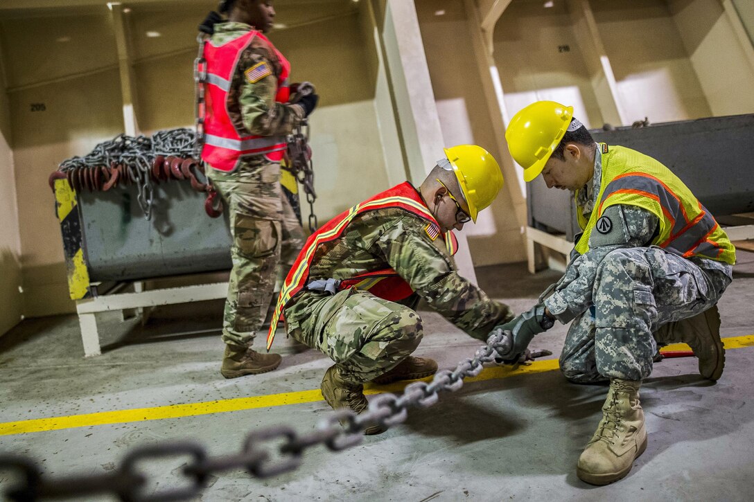 Two soldiers in hard hats and vests fasten a chain to an asphalt floor.