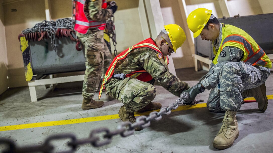 Two soldiers in hard hats and vests fasten a chain to an asphalt floor.