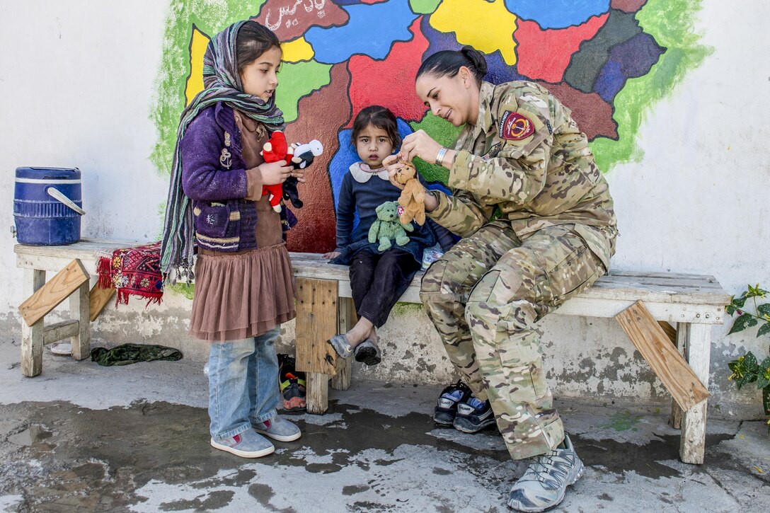 A soldier and two girls handle small stuffed animals while gathered in front of a bright mural.