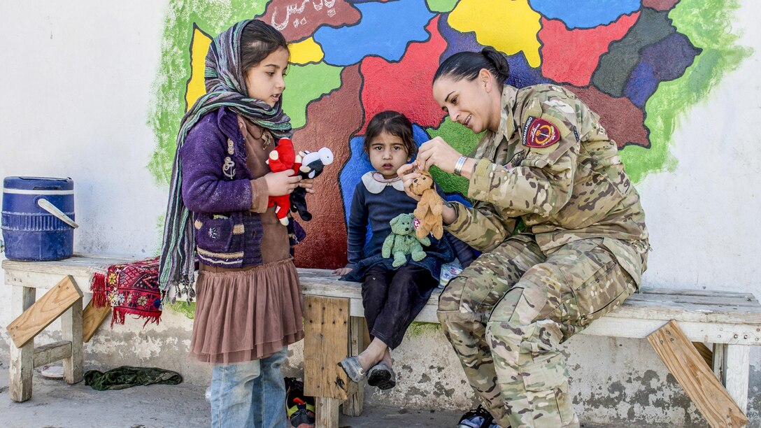 A soldier and two girls handle small stuffed animals while gathered in front of a bright mural.
