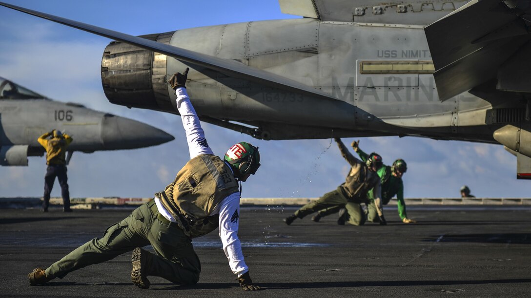 Three Marines kneeling on a flight deck hold their thumbs up on either side of an aircraft.