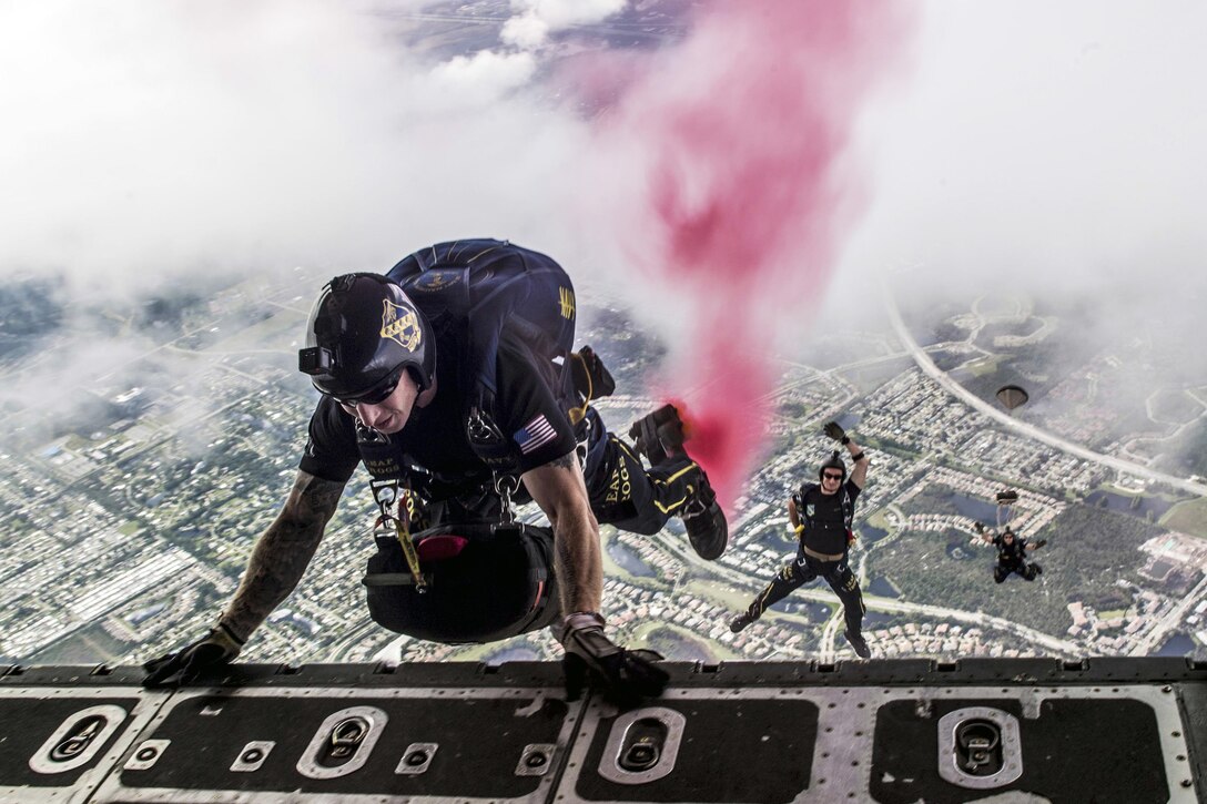 One parachutist holds onto the open door of an aircraft as two others fall in the distance below him.