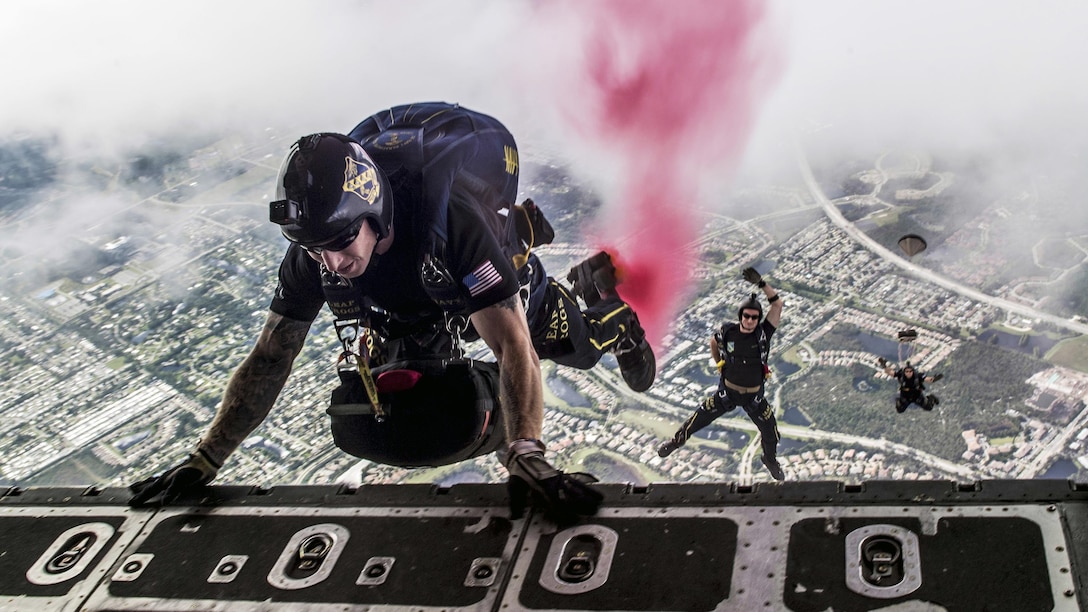 One parachutist holds onto the open door of an aircraft as two others fall in the distance below him.