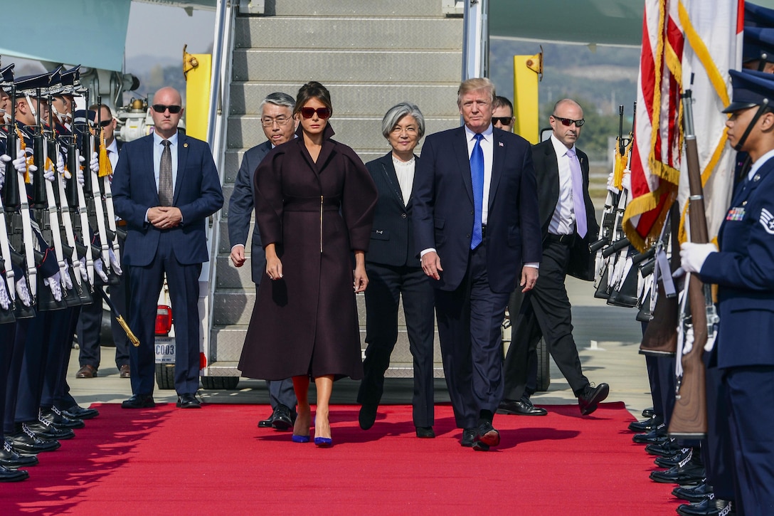 President Donald J. Trump and First Lady Melania Trump walk on a red carpet on a flightline, with rows of troops on either side.