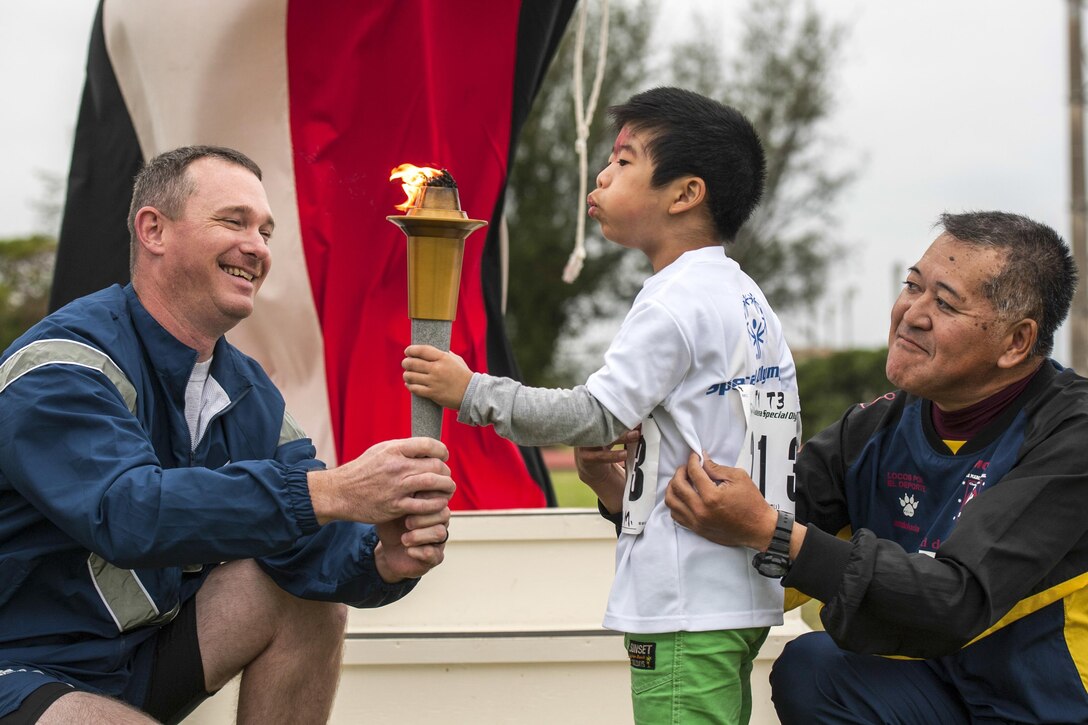 An airman helps a child holds a torch, as the child, held by his father, blows its flame.