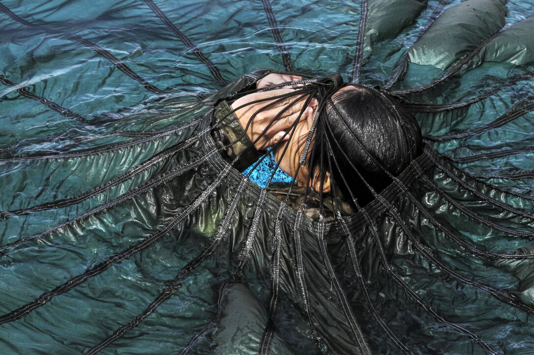A solider in water holds her hands in front of her face with a parachute over her.