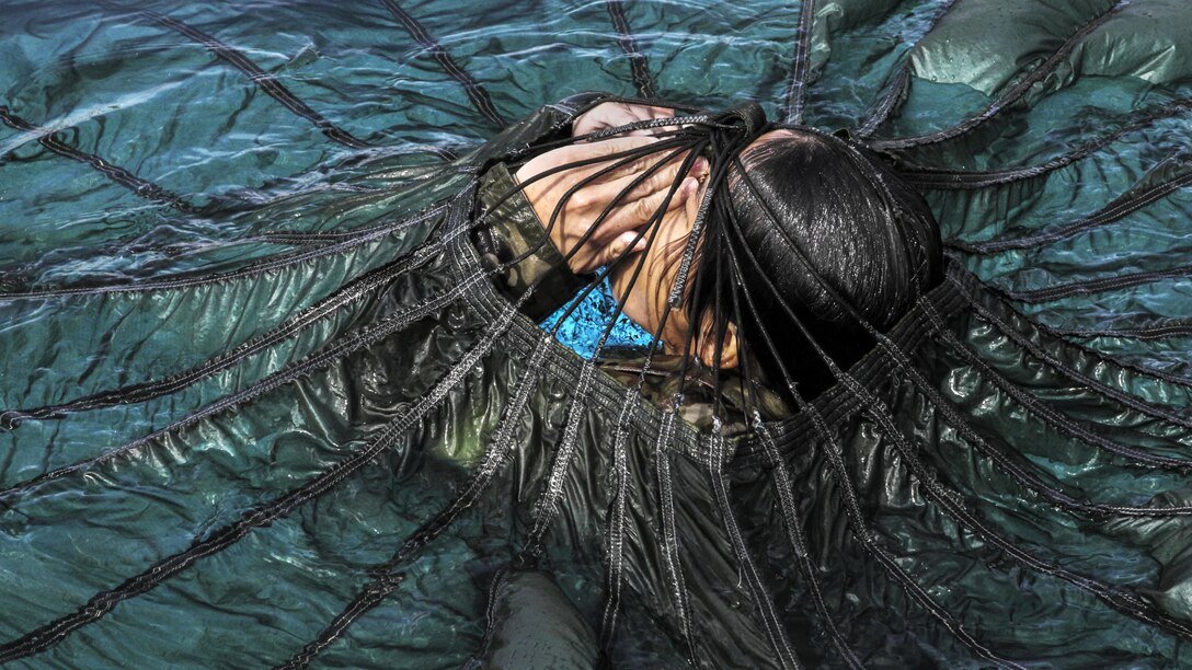 A solider in water holds her hands in front of her face with a parachute over her.