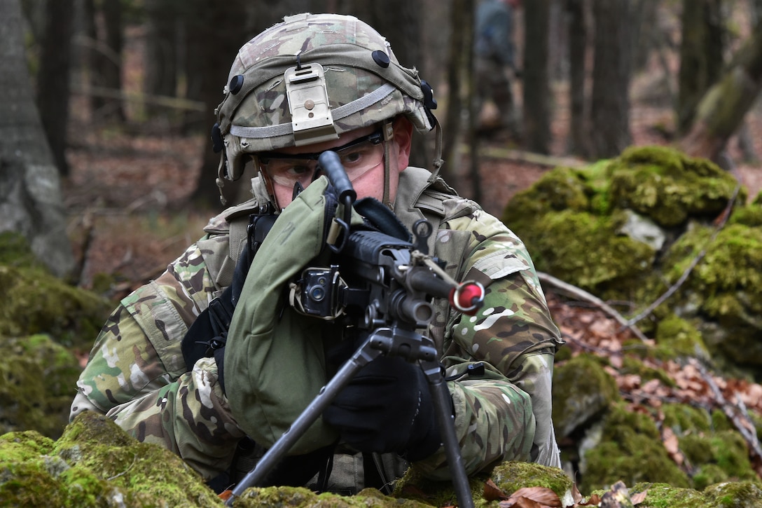 A soldier lays on the ground in a forest and aims a firearm.