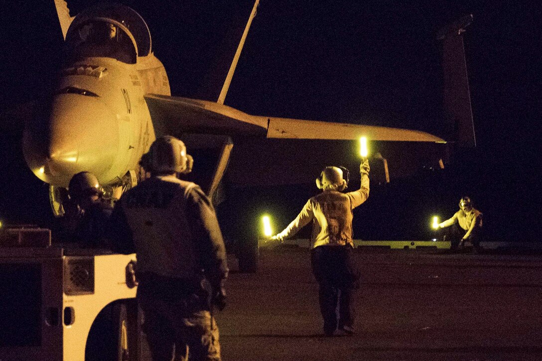 Sailors guide a fighter jet during night flight deck operations on an aircraft carrier.