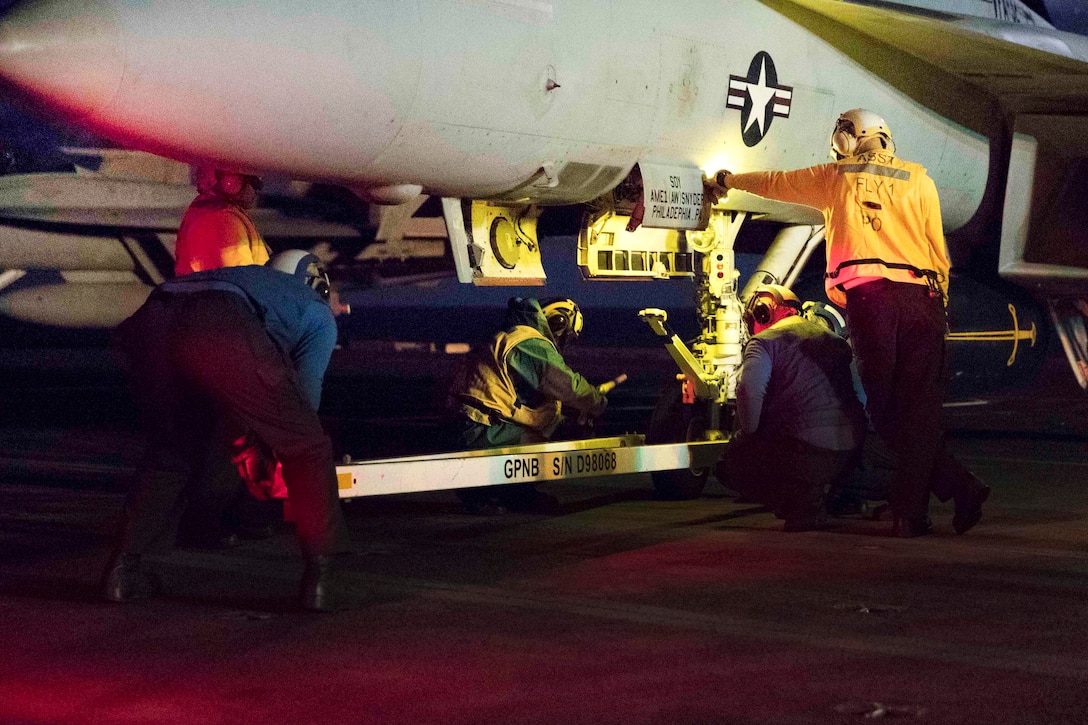 Sailors hook up a tow bar to a fighter jet aboard an aircraft carrier at night.