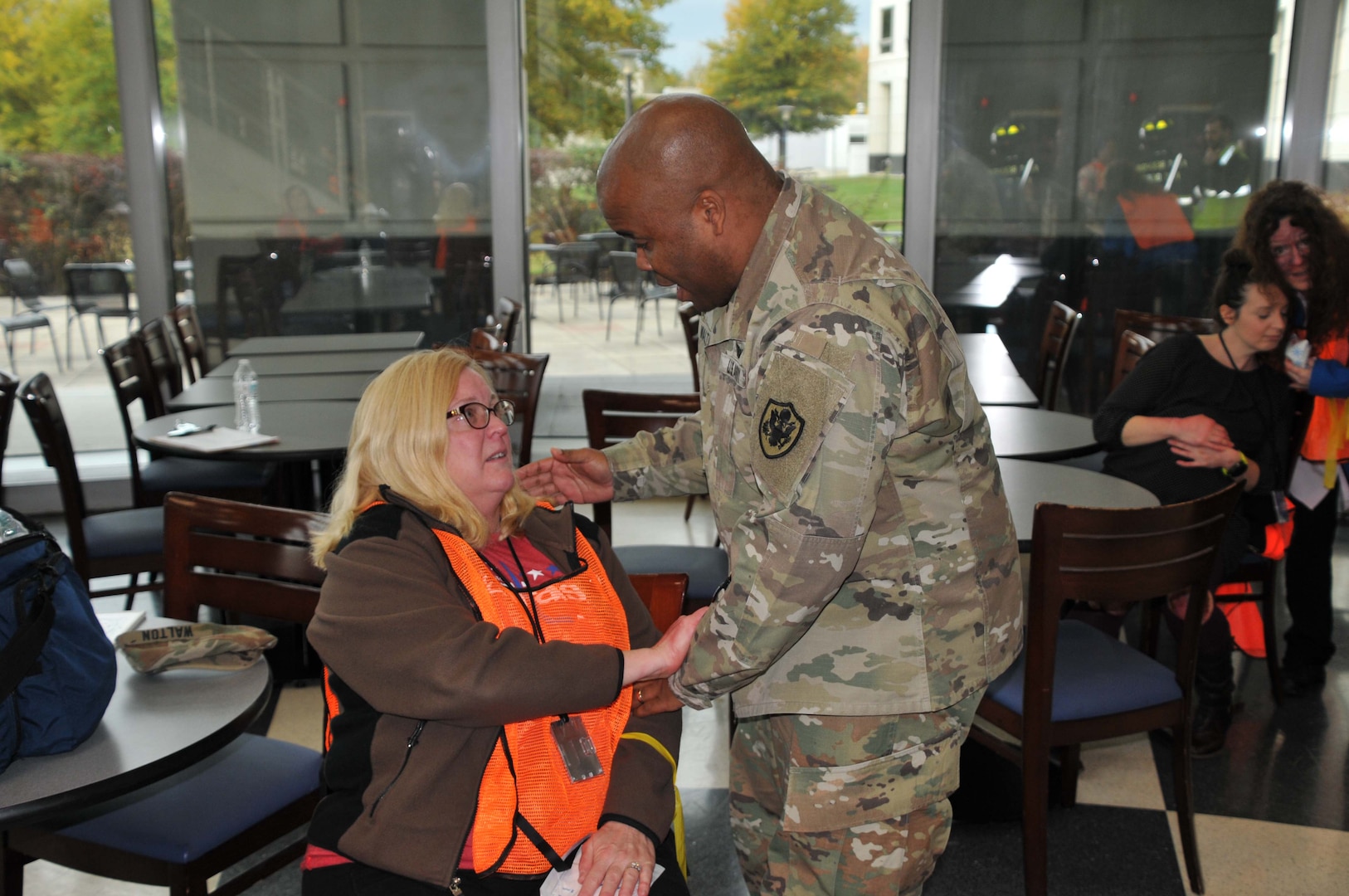 Chaplain in fatigues standing, speaking with 'victim', seated.