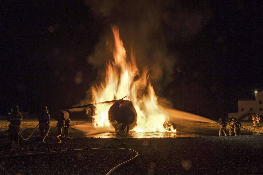 Firefighters on either side of an aircraft engulfed in flames spray water on it at night.