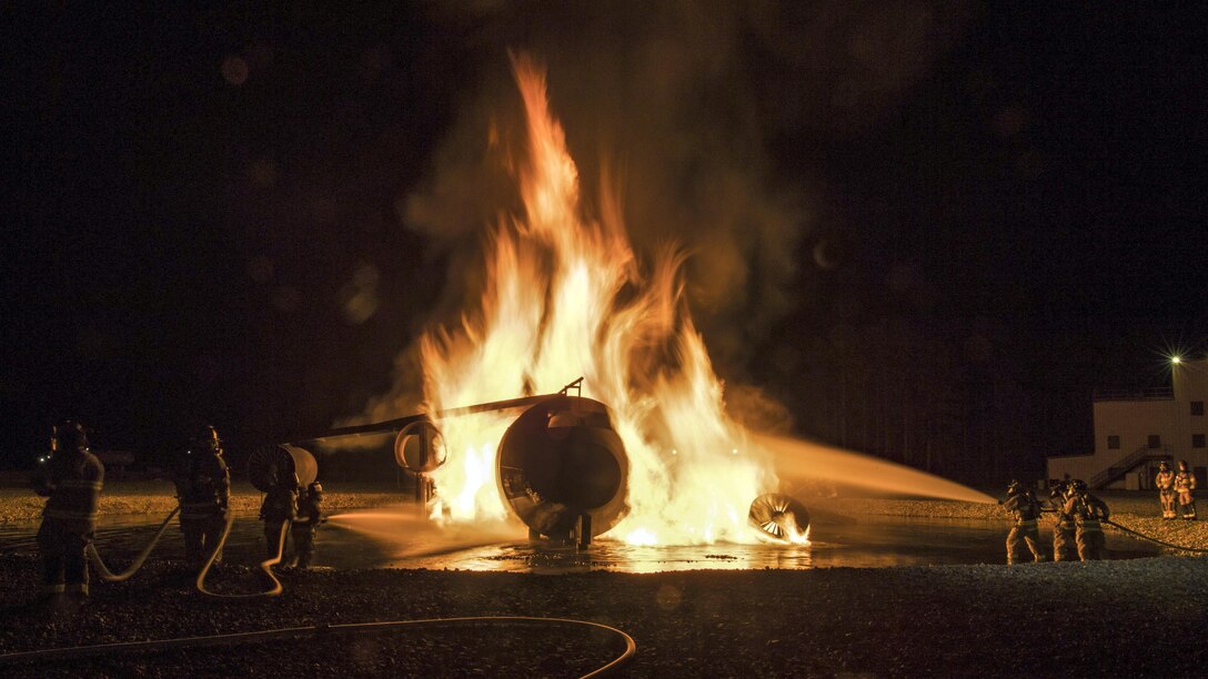 Firefighters on either side of an aircraft engulfed in flames spray water on it at night.