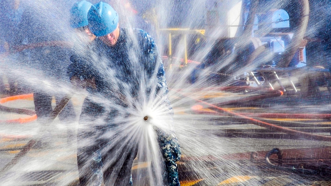 Water from a spraying hose held by two sailors creates a starburst effect.