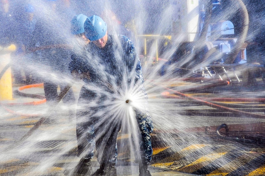 Water from a spraying hose held by two sailors creates a starburst effect.