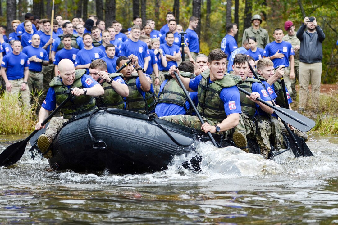 Soldiers in an inflatable boat paddle across a lake as fellow troops watch from the shore.