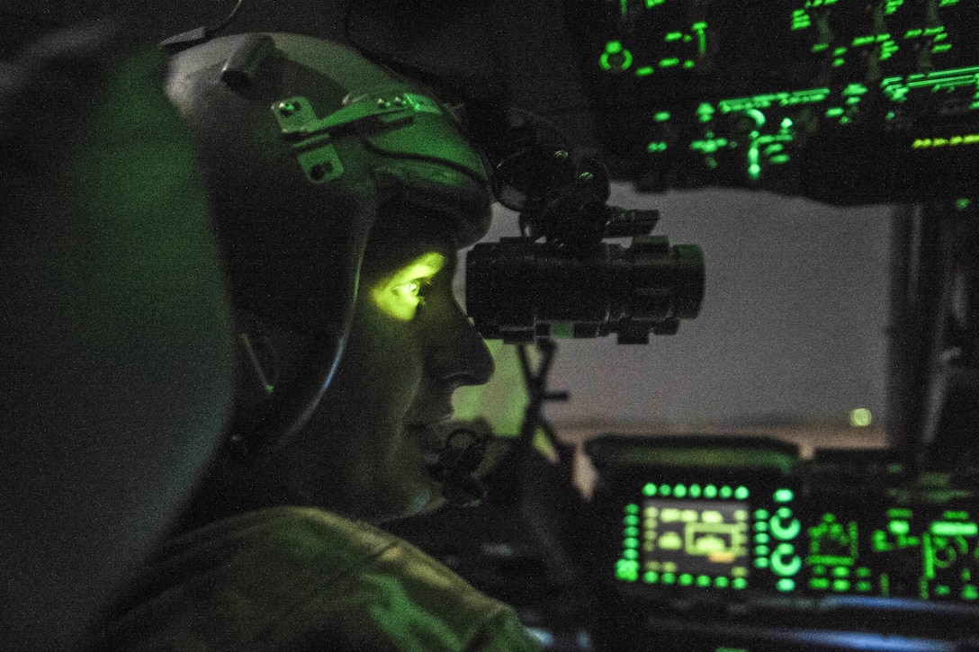 A pilot, shown in profile, looks through a scope in a cockpit, as green light illuminates his eye.
