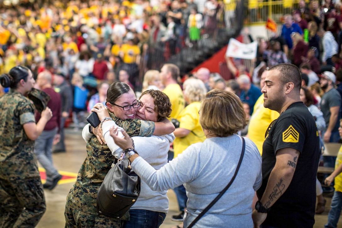 A smiling Marine hugs a female relative, amid of a crowd of Marines and civilians.