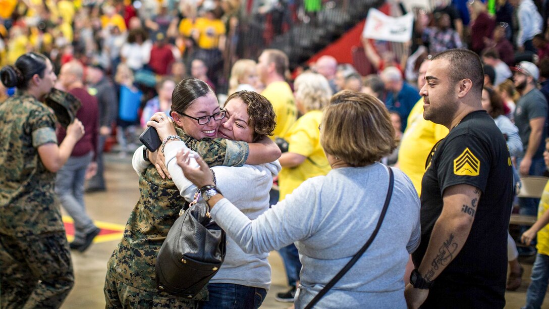 A smiling Marine hugs a female relative, amid of a crowd of Marines and civilians.