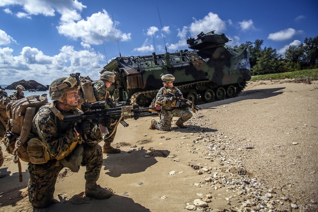 Marines kneel on a beach with weapons near an amphibious vehicle.