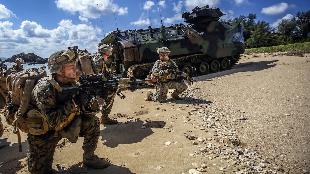 Marines kneel on a beach with weapons near an amphibious vehicle.