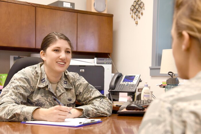Two service members sit at a desk and speak to each other.