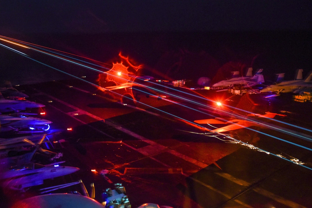 An aircraft lands on an aircraft carrier at night as red and blue lights glow.