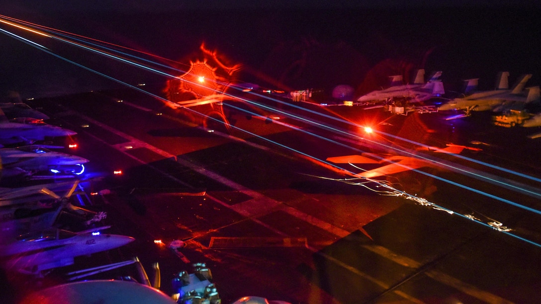 An aircraft lands on an aircraft carrier at night as red and blue lights glow.