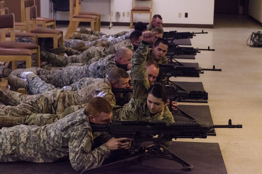 A soldier raises her hand while laying next to a gunner.