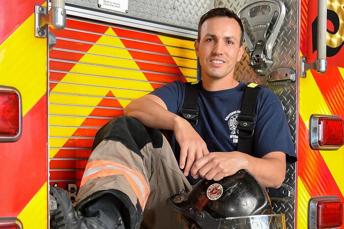 An airman sits on the back of a firetruck with firefighting gear.