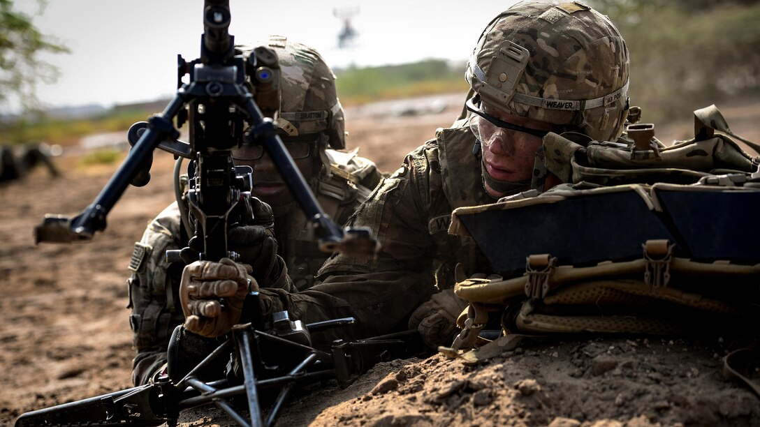 Two soldiers lay in sand about to fire a gun.