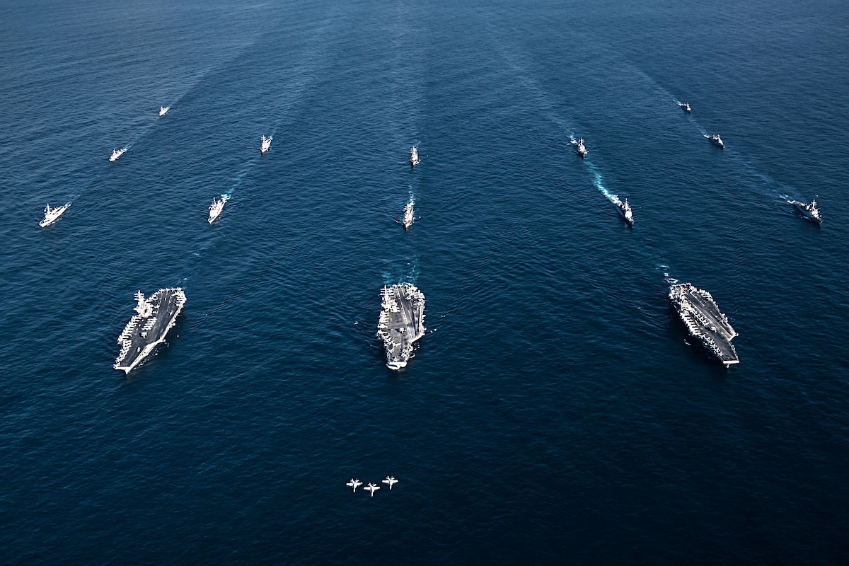 Three jets fly ahead of three carriers in formation in the ocean.