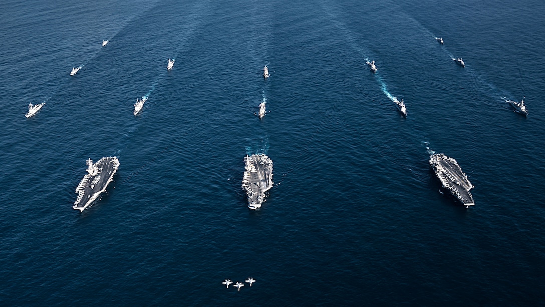 Three fighter aircraft fly in formation over three aircraft carriers in the ocean.