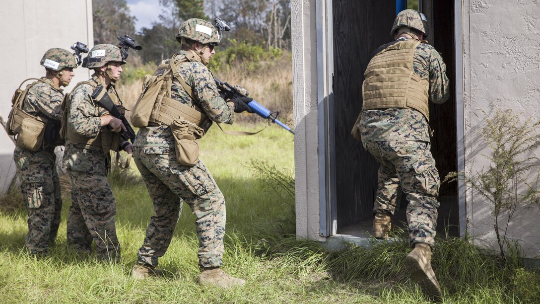 A Marine heads into a building as three others stand nearby wielding rifles.