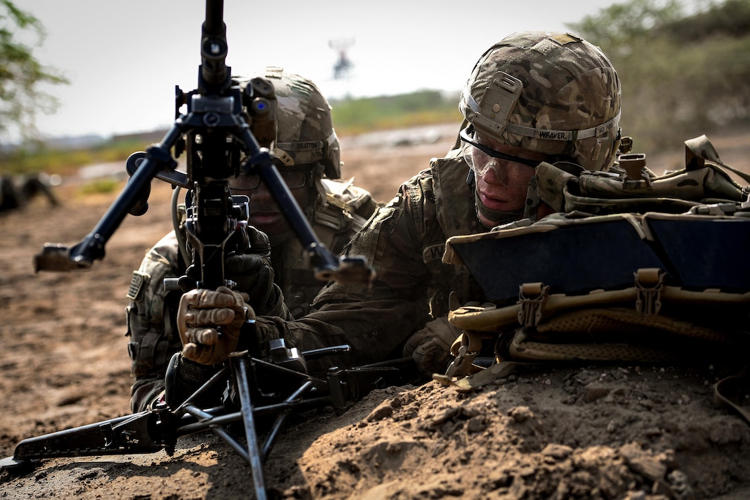 Two soldiers lay in sand about to fire a gun.