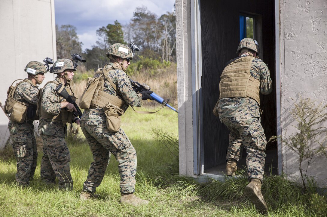 A Marine heads into a building as three others stand nearby wielding rifles.